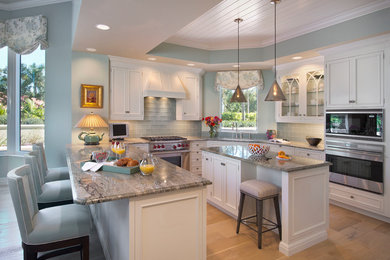 Example of a large beach style u-shaped light wood floor eat-in kitchen design in Miami with an undermount sink, recessed-panel cabinets, white cabinets, granite countertops, blue backsplash, glass tile backsplash, stainless steel appliances and an island