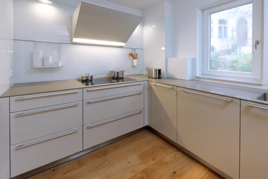 This is an example of a mid-sized contemporary u-shaped kitchen in Bremen with no island, white splashback, black appliances, a drop-in sink, flat-panel cabinets, white cabinets, light hardwood floors and brown floor.