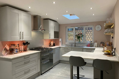 This is an example of a medium sized contemporary u-shaped enclosed kitchen in Surrey with shaker cabinets, grey cabinets, laminate countertops, metallic splashback, glass sheet splashback, stainless steel appliances, porcelain flooring, a breakfast bar, grey floors, white worktops and feature lighting.