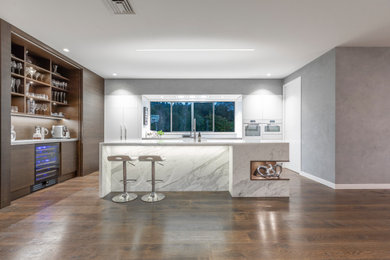 Inspiration for a large contemporary l-shaped eat-in kitchen in Auckland with a double-bowl sink, flat-panel cabinets, white cabinets, marble benchtops, white splashback, ceramic splashback, white appliances, medium hardwood floors, with island, brown floor and white benchtop.