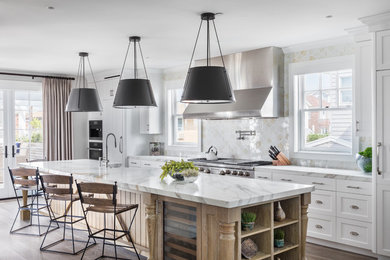 Example of a beach style galley medium tone wood floor and brown floor open concept kitchen design in New York with a farmhouse sink, recessed-panel cabinets, white cabinets, marble countertops, white backsplash, stainless steel appliances and an island