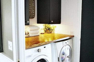 Imagen de lavadero de galera campestre pequeño con puertas de armario negras, encimera de madera, paredes blancas, suelo laminado y lavadora y secadora juntas