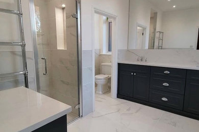 This is an example of a medium sized traditional ensuite bathroom in London with shaker cabinets, blue cabinets, a walk-in shower, a one-piece toilet, white tiles, stone tiles, a built-in sink, marble worktops, white floors, a hinged door, white worktops, double sinks and a built in vanity unit.