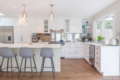 Photo of a large transitional l-shaped eat-in kitchen in Auckland with an undermount sink, shaker cabinets, white cabinets, mirror splashback, stainless steel appliances, medium hardwood floors, with island, brown floor and white benchtop.