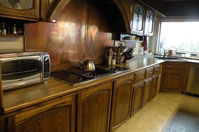 This is an example of a large traditional l-shaped eat-in kitchen in Seattle with a drop-in sink, raised-panel cabinets, medium wood cabinets, wood benchtops, metallic splashback, travertine floors, with island and black appliances.