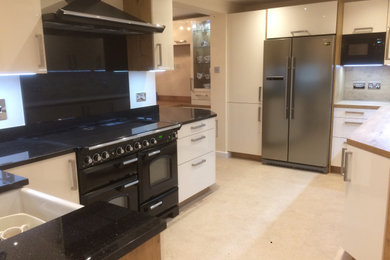 This is an example of a large traditional eat-in kitchen in Cambridgeshire with a farmhouse sink, flat-panel cabinets, white cabinets, granite benchtops, brown splashback, timber splashback, black appliances, no island and black benchtop.