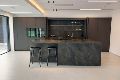 Photo of a large modern galley open plan kitchen in Hertfordshire with a drop-in sink, flat-panel cabinets, dark wood cabinets, quartzite benchtops, brown splashback, engineered quartz splashback, black appliances, concrete floors, with island, grey floor, brown benchtop and recessed.