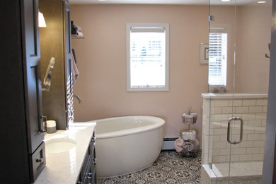 Photo of a mid-sized transitional master bathroom in Providence with recessed-panel cabinets, brown cabinets, a freestanding tub, an alcove shower, beige tile, subway tile, pink walls, cement tiles, an undermount sink, engineered quartz benchtops, white floor, a hinged shower door and white benchtops.