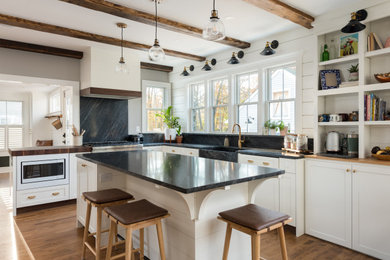 Inspiration for a mid-sized country l-shaped medium tone wood floor and exposed beam open concept kitchen remodel in Boston with a farmhouse sink, shaker cabinets, white cabinets, granite countertops, black backsplash, granite backsplash, stainless steel appliances, an island and black countertops