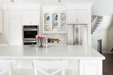Mid-sized transitional l-shaped dark wood floor and brown floor open concept kitchen photo in Toronto with an undermount sink, recessed-panel cabinets, white cabinets, marble countertops, white backsplash, ceramic backsplash, stainless steel appliances and an island