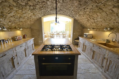 Immagine di un cucina con isola centrale country con lavello integrato, ante con riquadro incassato, ante con finitura invecchiata, top in legno, paraspruzzi in travertino, elettrodomestici neri e pavimento in pietra calcarea