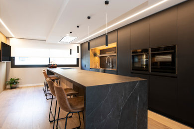 Inspiration for a large modern galley open plan kitchen in Barcelona with a submerged sink, flat-panel cabinets, black cabinets, engineered stone countertops, black splashback, engineered quartz splashback, black appliances, laminate floors, an island, brown floors and black worktops.
