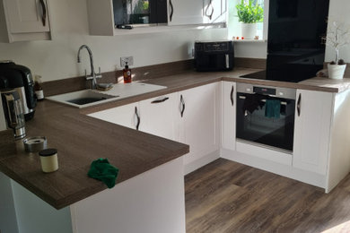 This is an example of a small contemporary u-shaped enclosed kitchen in West Midlands with a double-bowl sink, shaker cabinets, white cabinets, laminate countertops, black splashback, glass sheet splashback, black appliances, laminate floors, no island, brown floors, brown worktops and a coffered ceiling.