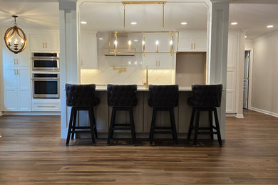 Example of a minimalist single-wall medium tone wood floor and brown floor eat-in kitchen design in Other with shaker cabinets, white cabinets, quartz countertops, white backsplash, ceramic backsplash, stainless steel appliances, an island and white countertops