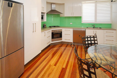 Mid-sized modern l-shaped eat-in kitchen in Adelaide with a double-bowl sink, flat-panel cabinets, white cabinets, laminate benchtops, green splashback, stainless steel appliances, medium hardwood floors and no island.