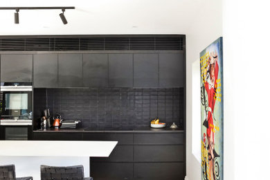 Photo of a contemporary galley kitchen in Sydney with flat-panel cabinets, black splashback, stainless steel appliances, light hardwood floors, with island, beige floor and black benchtop.