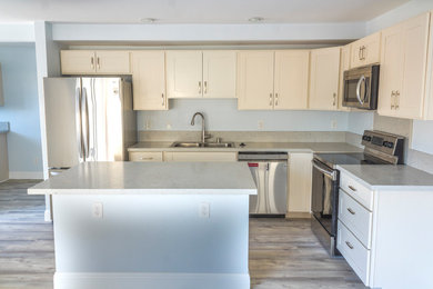 Photo of a l-shaped kitchen in Hawaii with a double-bowl sink, shaker cabinets, white cabinets, composite countertops, stainless steel appliances, vinyl flooring, an island, grey floors and white worktops.