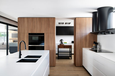 Exemple d'une grande arrière-cuisine tendance en L avec un évier encastré, un placard à porte plane, un plan de travail en surface solide, une crédence blanche, un électroménager noir, îlot, un plan de travail blanc et un sol en bois brun.