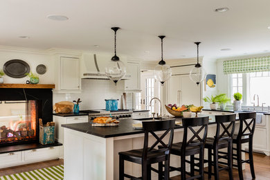 Large transitional l-shaped medium tone wood floor and brown floor eat-in kitchen photo in Boston with a farmhouse sink, flat-panel cabinets, white cabinets, soapstone countertops, white backsplash, subway tile backsplash, paneled appliances and an island