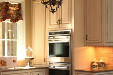 Eat-in kitchen - large traditional eat-in kitchen idea in Other with raised-panel cabinets, distressed cabinets, granite countertops, beige backsplash, stone tile backsplash, stainless steel appliances and an island
