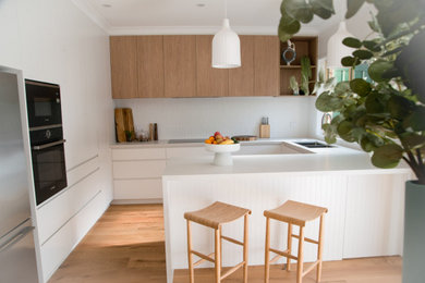 Inspiration for a mid-sized modern u-shaped eat-in kitchen in Sydney with a double-bowl sink, flat-panel cabinets, white cabinets, quartz benchtops, white splashback, subway tile splashback, black appliances, laminate floors, a peninsula, brown floor and white benchtop.