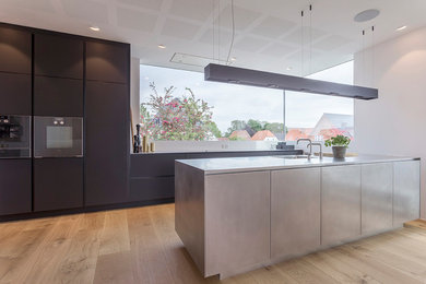 This is an example of a large galley eat-in kitchen in Esbjerg with an integrated sink, flat-panel cabinets, black cabinets, stainless steel benchtops, light hardwood floors and with island.