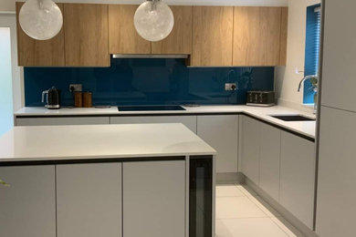 Design ideas for a medium sized contemporary grey and white l-shaped kitchen with an integrated sink, flat-panel cabinets, grey cabinets, quartz worktops, blue splashback, glass sheet splashback, integrated appliances, ceramic flooring, an island, white worktops and feature lighting.