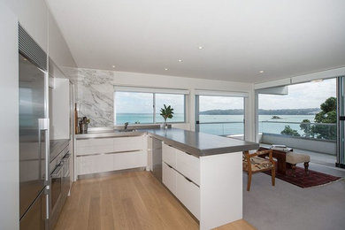 This is an example of a mid-sized contemporary u-shaped kitchen in Auckland with a drop-in sink, flat-panel cabinets, white cabinets, stainless steel benchtops, multi-coloured splashback, stone slab splashback, stainless steel appliances and light hardwood floors.