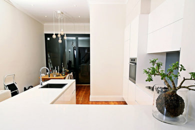 This is an example of a contemporary u-shaped eat-in kitchen in Sydney with a single-bowl sink, flat-panel cabinets, white cabinets, granite benchtops, blue splashback, glass sheet splashback, stainless steel appliances, medium hardwood floors and no island.