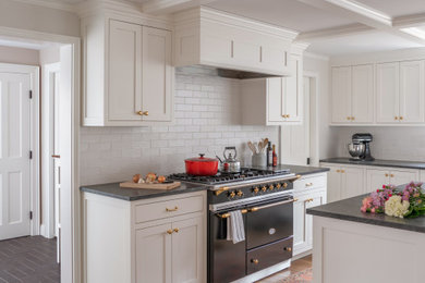 This is an example of a mid-sized traditional l-shaped eat-in kitchen in Boston with with island, a farmhouse sink, raised-panel cabinets, white cabinets, white splashback, subway tile splashback, black appliances, medium hardwood floors, brown floor and black benchtop.