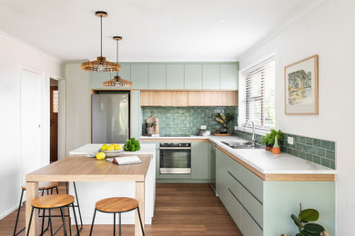 This is an example of a contemporary l-shaped kitchen in Hobart with a drop-in sink, flat-panel cabinets, green cabinets, green splashback, subway tile splashback, stainless steel appliances, medium hardwood floors, with island, brown floor and white benchtop.