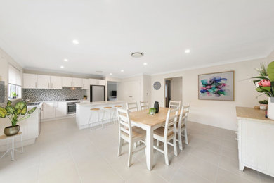 Inspiration for a large beach style l-shaped eat-in kitchen in Newcastle - Maitland with flat-panel cabinets, white cabinets, quartz benchtops, multi-coloured splashback, glass tile splashback, ceramic floors, with island, beige floor and white benchtop.