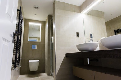This is an example of a medium sized contemporary ensuite bathroom in London with flat-panel cabinets, dark wood cabinets, a built-in shower, a wall mounted toilet, beige tiles, porcelain tiles, beige walls, porcelain flooring, a vessel sink, laminate worktops, beige floors, a hinged door and brown worktops.