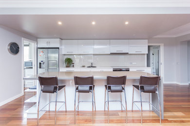 This is an example of a mid-sized contemporary single-wall eat-in kitchen in Melbourne with flat-panel cabinets, white cabinets, quartz benchtops, beige splashback, glass sheet splashback, stainless steel appliances, medium hardwood floors, with island, brown floor and white benchtop.