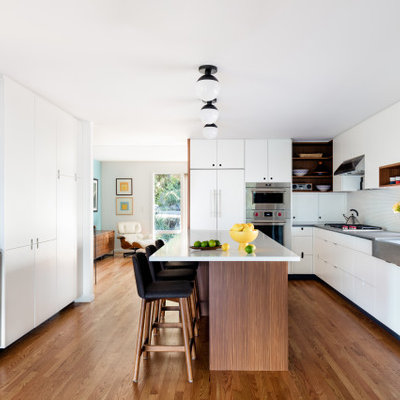 Eat-in kitchen - mid-sized mid-century modern l-shaped medium tone wood floor and brown floor eat-in kitchen idea in Seattle with flat-panel cabinets, white cabinets, solid surface countertops, stainless steel appliances, an island, white countertops, a farmhouse sink, white backsplash and mosaic tile backsplash