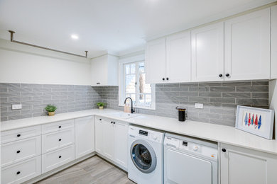 This is an example of a large contemporary u-shaped dedicated laundry room in Brisbane with shaker cabinets, white cabinets, quartz benchtops, grey splashback, subway tile splashback, porcelain floors, grey floor, white benchtop, a drop-in sink, white walls and a side-by-side washer and dryer.