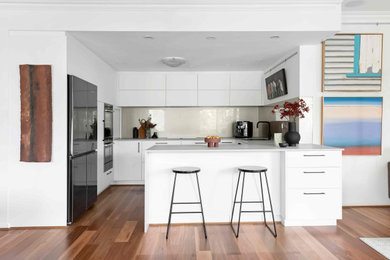 Photo of a mid-sized modern galley open plan kitchen in Perth with a drop-in sink, recessed-panel cabinets, white cabinets, quartz benchtops, grey splashback, glass sheet splashback, black appliances, medium hardwood floors, no island, brown floor and grey benchtop.