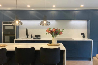 This is an example of a large contemporary kitchen/diner with shaker cabinets, blue cabinets, quartz worktops, white splashback, engineered quartz splashback, integrated appliances, vinyl flooring, an island, brown floors, white worktops and feature lighting.