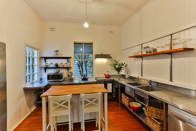 Small industrial l-shaped open plan kitchen in Brisbane with a double-bowl sink, open cabinets, stainless steel cabinets, stainless steel benchtops, white splashback, stainless steel appliances, medium hardwood floors and with island.