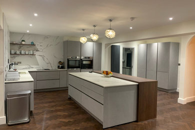 Inspiration for a medium sized modern l-shaped kitchen/diner in Cheshire with flat-panel cabinets, white cabinets, engineered stone countertops, black appliances, an island, white worktops, a built-in sink, dark hardwood flooring, brown floors and feature lighting.