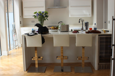Inspiration for a medium sized victorian l-shaped kitchen/diner in Surrey with an integrated sink, flat-panel cabinets, white cabinets, composite countertops, beige splashback, glass sheet splashback, stainless steel appliances, light hardwood flooring, an island, brown floors, beige worktops, a vaulted ceiling and feature lighting.