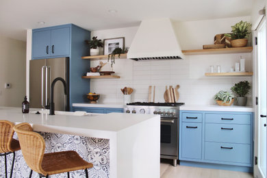 Example of a mid-sized minimalist galley light wood floor kitchen design with a farmhouse sink, flat-panel cabinets, blue cabinets, quartz countertops, white backsplash, porcelain backsplash, stainless steel appliances, an island and white countertops