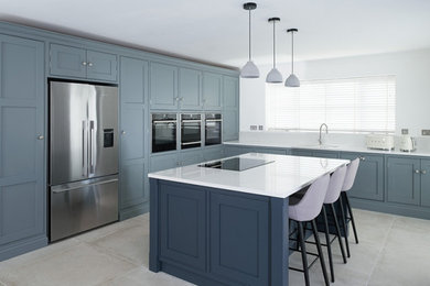This is an example of a large classic l-shaped kitchen/diner in Cheshire with a double-bowl sink, shaker cabinets, blue cabinets, quartz worktops, stainless steel appliances, limestone flooring, an island, beige floors and white worktops.