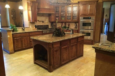 Example of a large ornate l-shaped light wood floor enclosed kitchen design in Dallas with an undermount sink, raised-panel cabinets, dark wood cabinets, granite countertops, beige backsplash, stone tile backsplash, stainless steel appliances and an island