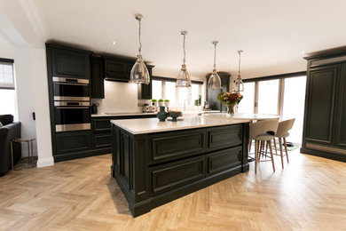 Inspiration for a large traditional galley open plan kitchen in Essex with a double-bowl sink, shaker cabinets, green cabinets, quartz worktops, white splashback, engineered quartz splashback, stainless steel appliances, light hardwood flooring, an island, beige floors, white worktops and a feature wall.