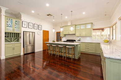 Large country l-shaped kitchen in Perth with a farmhouse sink, glass-front cabinets, green cabinets, marble benchtops, white splashback, subway tile splashback, white appliances, dark hardwood floors, with island, brown floor, multi-coloured benchtop and exposed beam.