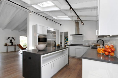 Photo of a mid-sized contemporary l-shaped open plan kitchen in Los Angeles with an undermount sink, flat-panel cabinets, white cabinets, grey splashback, stainless steel appliances, medium hardwood floors, with island, soapstone benchtops and ceramic splashback.