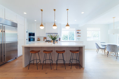 Inspiration for a large contemporary l-shaped eat-in kitchen in Sacramento with a farmhouse sink, shaker cabinets, white cabinets, quartz benchtops, white splashback, subway tile splashback, stainless steel appliances, medium hardwood floors, with island, brown floor and white benchtop.