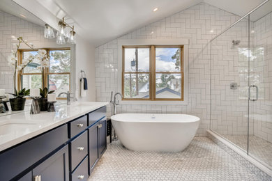 デンバーにあるトランジショナルスタイルのおしゃれな浴室 (シェーカースタイル扉のキャビネット、青いキャビネット、白いタイル、磁器タイル、白い壁、大理石の床、グレーの床、開き戸のシャワー、洗面台2つ、造り付け洗面台、置き型浴槽) の写真