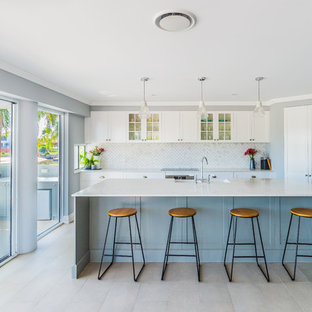 Beach style kitchen in Gold Coast - Tweed with glass-front cabinets, white cabinets, grey splashback, stainless steel appliances, with island, beige floor and grey benchtop.